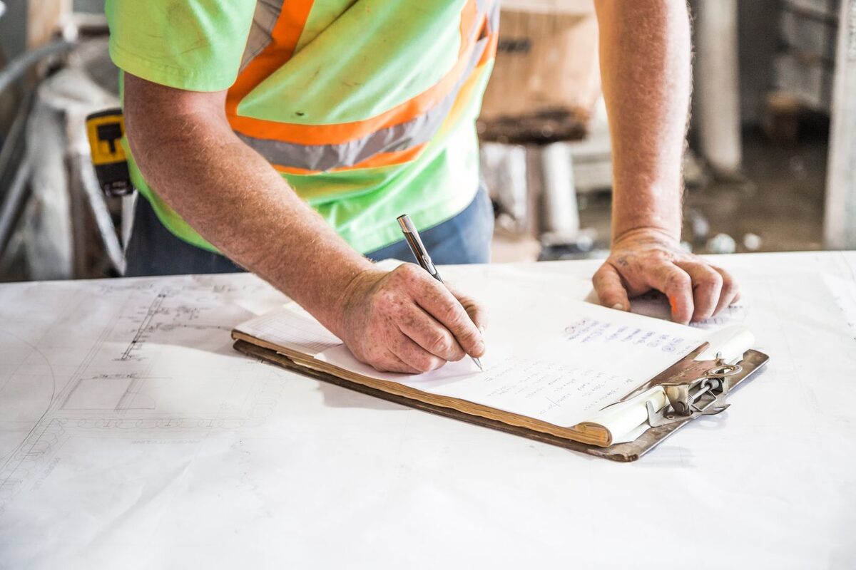 Worker in high-visibility gear writing notes on a clipboard over site drawings